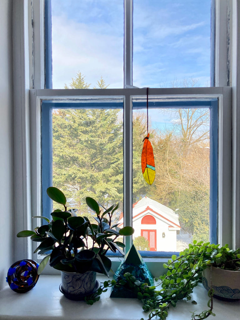 Stained glass feather with red, yellow, and blue panes hanging from leather tie in window