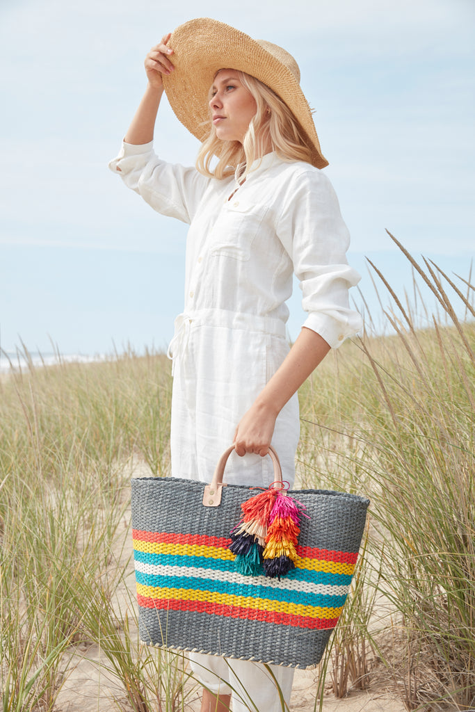 Model holding dove colored, multi-colored striped basket tote with removable tassels and leather handles