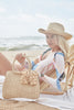 Model sitting on a beach chair wearing our Rose raffia sun hat in Natural and our raffia Lauren handbag in Natural