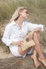 Model sitting on beachy driftwood holding our Sofia raffia clutch in Multi