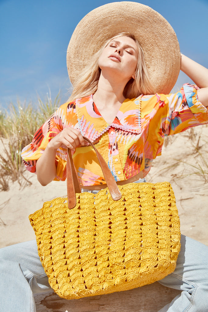 Model sitting on a beach wearing our crocheted raffia Jane hat in Natural and crocheted raffia Paros Tote in Sunflower with a colorful shirt and jeans