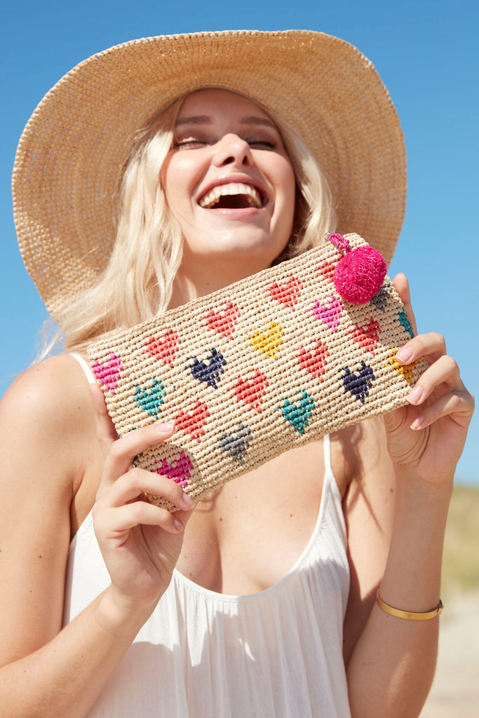 Model holding multi colored crocheted clutch with a heart pattern, raffia pom poms and zip closure.