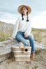 Model sitting on driftwood  wearing our Mika hat in Natural and our Capri tote in Sand sitting by her feet