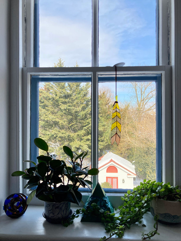 Stained glass feather with grey, yellow, and pink panes hanging from leather tie in window
