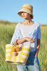 Model holding sunflower colored striped woven sisal basket tote with pom poms and leather handles