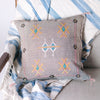 Dove colored square pillow with embroidered design on a blanket and chair