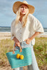 Model on the beach wearing our Mika raffia sun hat in Natural and our woven sisal Hadley tote in Aqua