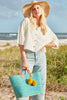 Model on the beach wearing our Mika raffia sun hat in Natural and our Hadley beach tote in Aqua