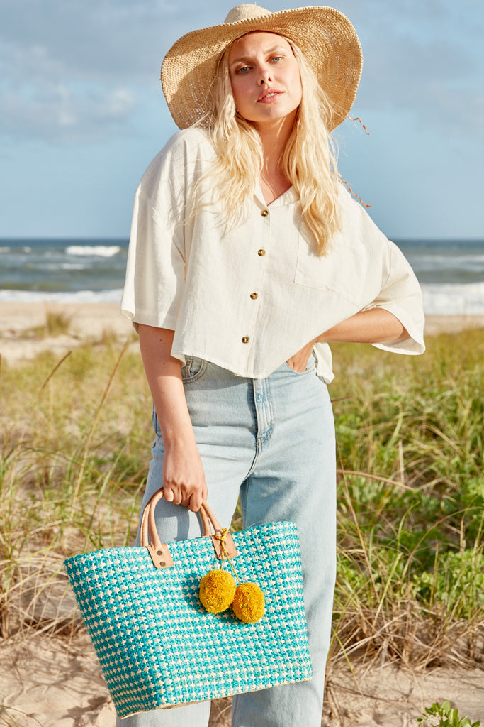 Model on the beach wearing our Mika raffia sun hat in Natural and our Hadley beach tote in Aqua