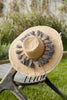 Wide brim braided raffia sun hat with a ring of dove colored tassels on a chair