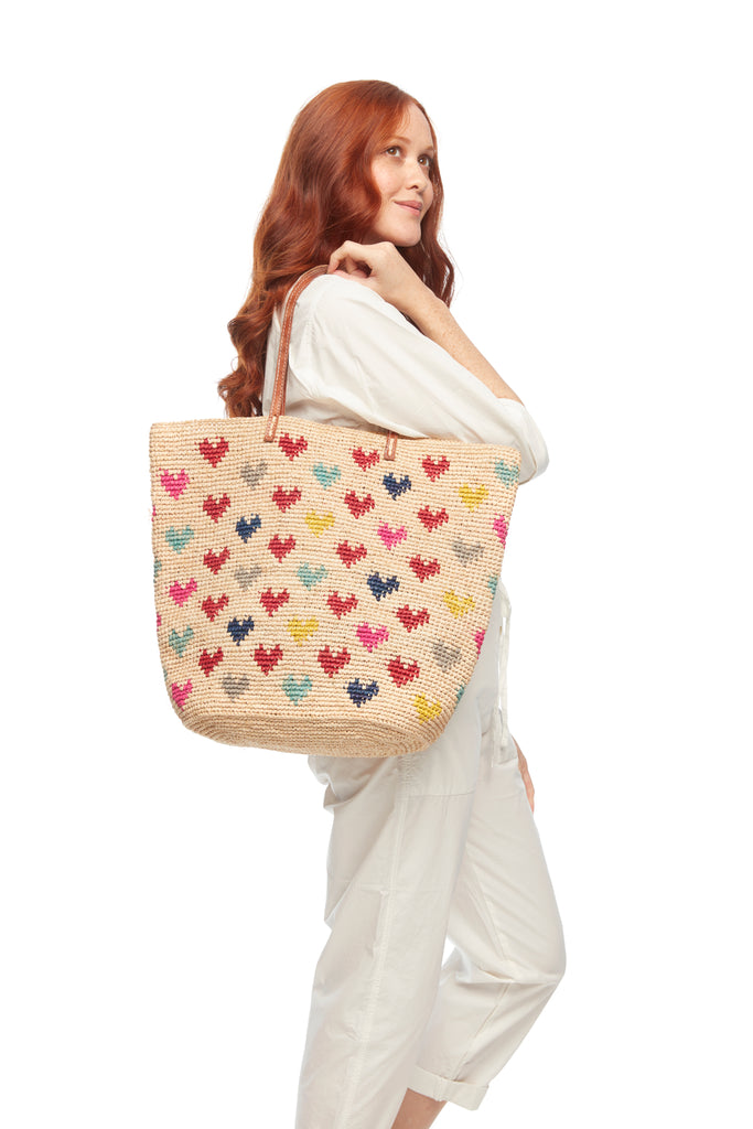 Model holding crocheted carryall with multi colored heart pattern, cotton lining, snap closure, & leather straps