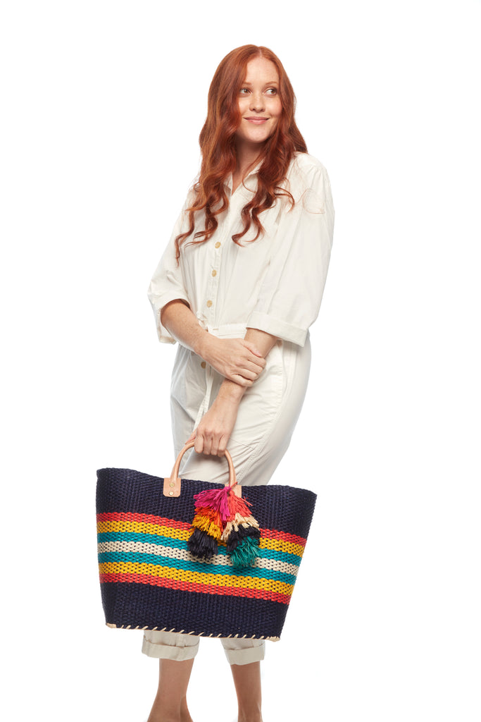 Model holding navy colored, multi-colored striped basket tote with removable tassels and leather handles