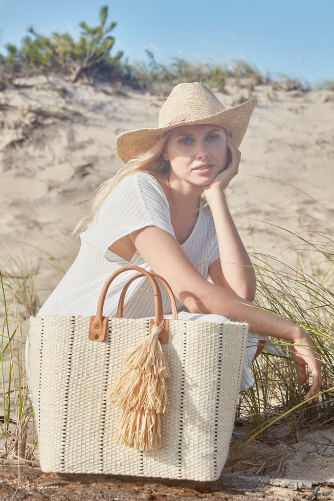 Model on the beach wearing our Mika raffia sun hat in Natural and our Marley sisal beach tote in Natural