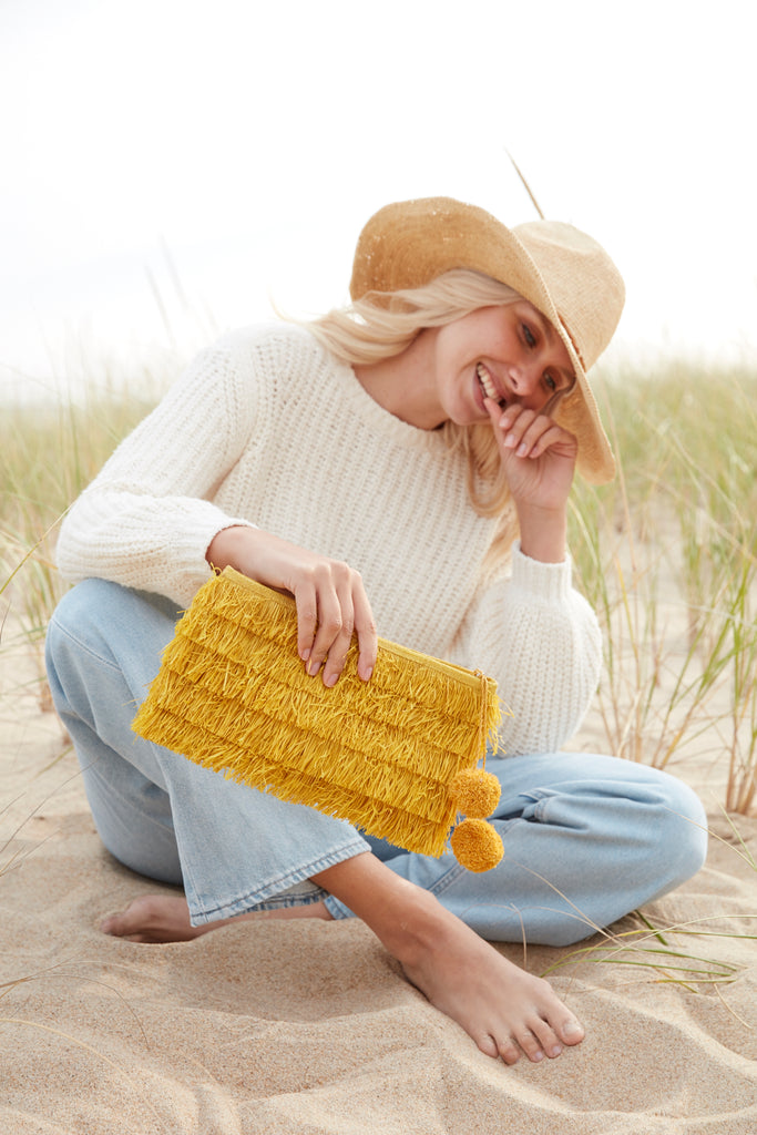 Model holding sunflower colored woven raffia fringe clutch with pom poms and zip closure