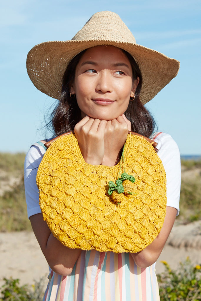 Model holding sunflower colored crocheted raffia clutch with wooden handles, cotton lining, and pineapple charms